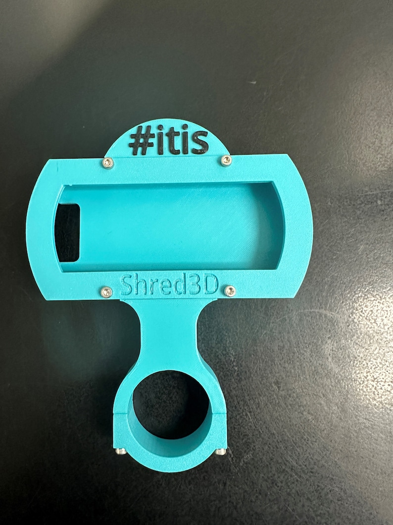 Peut inclure: Un support de t&eacute;l&eacute;phone imprim&eacute; en 3D bleu turquoise avec un collier de serrage pour guidon. Le support a une fente pour un t&eacute;l&eacute;phone et le texte "#itis" et "Shred3D" imprim&eacute; dessus.