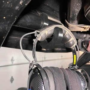 May include: Black headphones with a carbon fiber finish hanging from a black hook. The headphones have a yellow label with the word "Rugged" on it.
