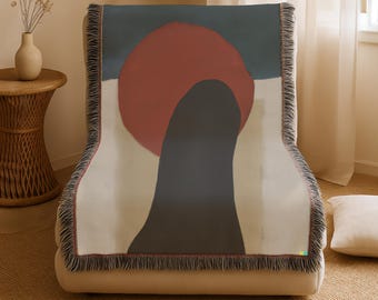 May include: A woven throw blanket with an abstract design. The artwork features a large red circle, a dark gray shape, and a blue-gray background. The blanket has a fringed edge and is draped over a beige chair.