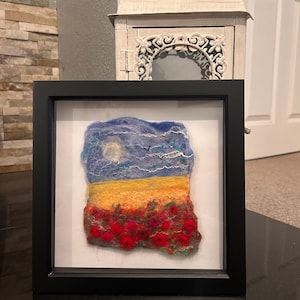May include: A square black frame containing a needle felted landscape art piece. The scene features a blue sky with a white moon and clouds, a yellow field, and a foreground filled with vibrant red poppy flowers on a textured fibre surface.