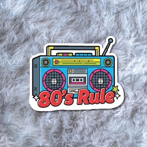Puede incluir: Una pegatina con un diseño de radiocasete retro. El radiocasete es principalmente azul con detalles rosas y amarillos, y una ranura para casete. El texto "80's Rule" es rojo con contorno blanco. Una estrella a la derecha.