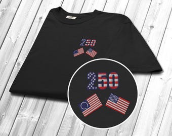 May include: Black t-shirt with embroidered design. Features the number "250" and two American flags. The "250" is in red, white, and blue. The flags are red, white, and blue. The t-shirt is on a light wood grain surface.
