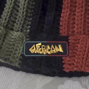 May include: A close-up of a knitted beanie in a color block design. The beanie features sections of olive green, black, and red. A rectangular patch with the word "AMERICAN" in gold lettering is sewn on the front.