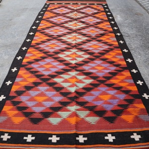 May include: A long, rectangular rug with a geometric pattern of black, red, orange, purple, and green triangles. The rug has a black border with white crosses.