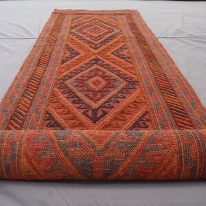 May include: A rolled-up red, orange, and gray patterned rug with a geometric design. The rug has a fringe border.