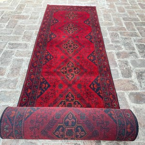 May include: A rolled-up red and navy blue patterned rug with a central diamond design. The rug is partially unrolled, revealing the intricate details of the woven pattern. The rug is placed on a brick surface.
