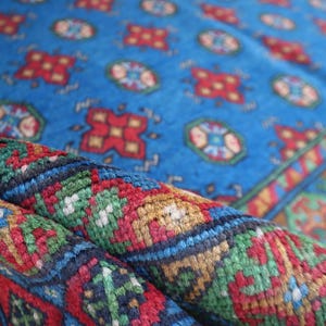 Puede incluir: Primer plano de una alfombra con patrones azules, rojos, verdes y dorados. La alfombra est&aacute; doblada por la mitad, mostrando los intrincados detalles del dise&ntilde;o tejido.