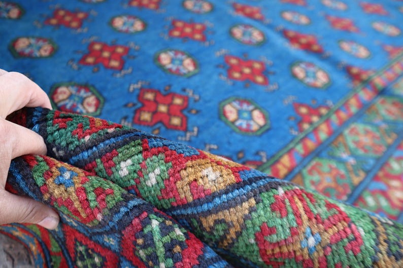 Puede incluir: Primer plano de una alfombra con patrones azules, rojos, verdes y dorados. La alfombra est&aacute; doblada por la mitad, mostrando los intrincados detalles del dise&ntilde;o tejido.