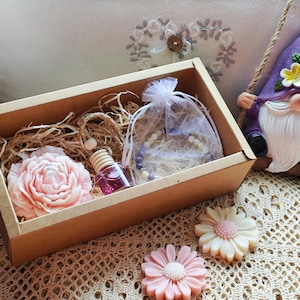 Può includere: Una confezione regalo con un sapone a forma di peonia rosa, un piccolo flacone di profumo e un braccialetto di perline. Sono visibili anche uno gnomo da giardino su un'altalena e due saponi a forma di fiore. La scatola è riempita di paglia.