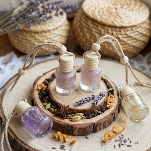 May include: Three small glass bottles with wooden caps and hanging jute cords, filled with shimmering purple and gold liquids. The bottles are arranged on a natural wood slice surrounded by dried lavender, star anise, cardamom, and orange peels.