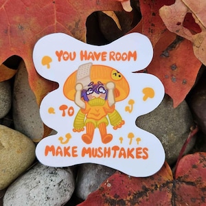 Könnte beinhalten: Ein weißer Aufkleber mit einer Cartoon-Pilzfigur und dem Text "YOU HAVE ROOM TO MAKE MUSHTAKES" in Orange. Der Aufkleber ist von bunten Herbstblättern und Steinen umgeben.