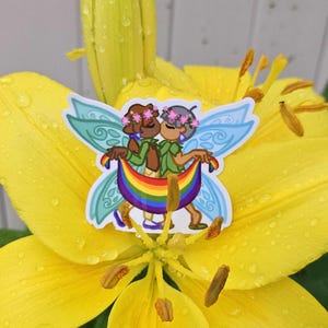 Könnte beinhalten: Ein Aufkleber mit zwei Cartoon-Figuren mit Feenflügeln, die sich umarmen und ein Regenbogenbanner halten. Der Aufkleber befindet sich auf einer leuchtend gelben Lilienblüte mit Wassertropfen. Die Figuren tragen Blumenkränze und grüne Kleidung.