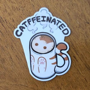Könnte beinhalten: Weißer Aufkleber mit einem Cartoon-Bild einer Katze in einer Kaffeetasse. Die Tasse hat braune und weiße Streifen und einen Pfotenabdruck. Der Text "CATFFEINATED" steht über der Tasse.