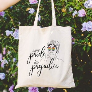 May include: A white tote bag with a black illustration of a woman wearing rainbow glasses and the text "more pride less prejudice".