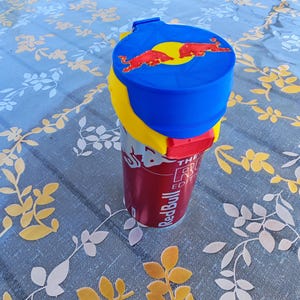Peut inclure: Une canette de boisson énergisante Red Bull au design rouge et blanc avec le texte Red Bull et The Red Edition. La canette est surmontée d'un couvercle en plastique bleu et jaune affichant deux taureaux rouges face à face sur un cercle solaire jaune.
