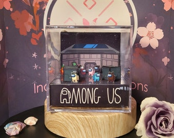 May include: A diorama featuring characters from the game Among Us, displayed in a clear acrylic box. The scene depicts the game's setting with crewmates and imposters. The base of the box has the game's title in white lettering.