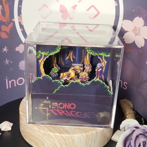May include: A clear acrylic cube display case featuring a 3D diorama of characters from the video game Chrono Trigger. The diorama depicts a forest scene with a group of characters gathered around a campfire. The base of the cube has a black background with the words "Chrono Trigger" printed in white.