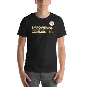 以下が含まれることがあります： 「EMPOWERING COMMUNITIES」のゴールドの文字がプリントされた濃いグレーのTシャツ。「PEOPLE FIRST ALWAYS」の文字が下にあります。Tシャツの右上には「SJ」の文字が入った白い丸があります。