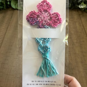 May include: A handmade crochet bookmark featuring a pink and purple flower design at the top and a turquoise tassel at the bottom. The bookmark is attached to a white card with a Bible verse printed on it. The bookmark is packaged in a clear plastic sleeve.