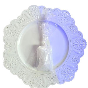 May include: A white, decorative plate with a scalloped edge featuring floral and dot patterns. A clear plastic bag is centered on the plate. The plate has a glossy finish and is set against a white background.