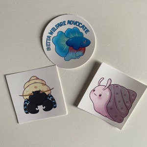 May include: Three stickers featuring cartoon animals. A blue betta fish with the text "Beta Welfare Advocate" in a circle. A hermit crab with a yellow shell and blue and black stripes. A pink snail with a purple shell.