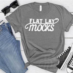 Pode incluir: Camiseta cinza com o texto "FLAT LAY MOCKS" em letras brancas. A camiseta está colocada sobre uma superfície de madeira branca com um par de calças de ganga azuis, óculos de sol e ténis pretos.