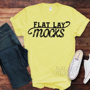 May include: A yellow t-shirt with the text "FLAT LAY MOCKS" in black lettering. The shirt is laid flat on a wooden surface with a pair of blue jeans and white sneakers.