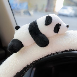 May include: A white plush steering wheel cover with a black and white panda plush toy attached to it. The panda is lying on the cover with its head resting on the steering wheel.