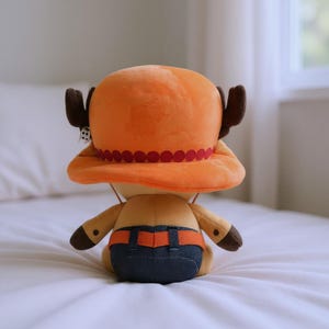 May include: A plush toy of a character from the anime series One Piece, seen from the back. The toy has an orange hat with a red trim, brown antlers, and a tan body with blue denim shorts. The toy is sitting on a white surface.