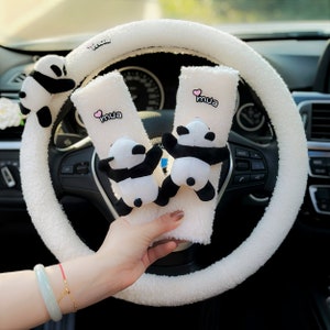 May include: White fuzzy steering wheel cover with two plush panda bear seatbelt covers. The seatbelt covers have the word "mua" embroidered on them in pink.