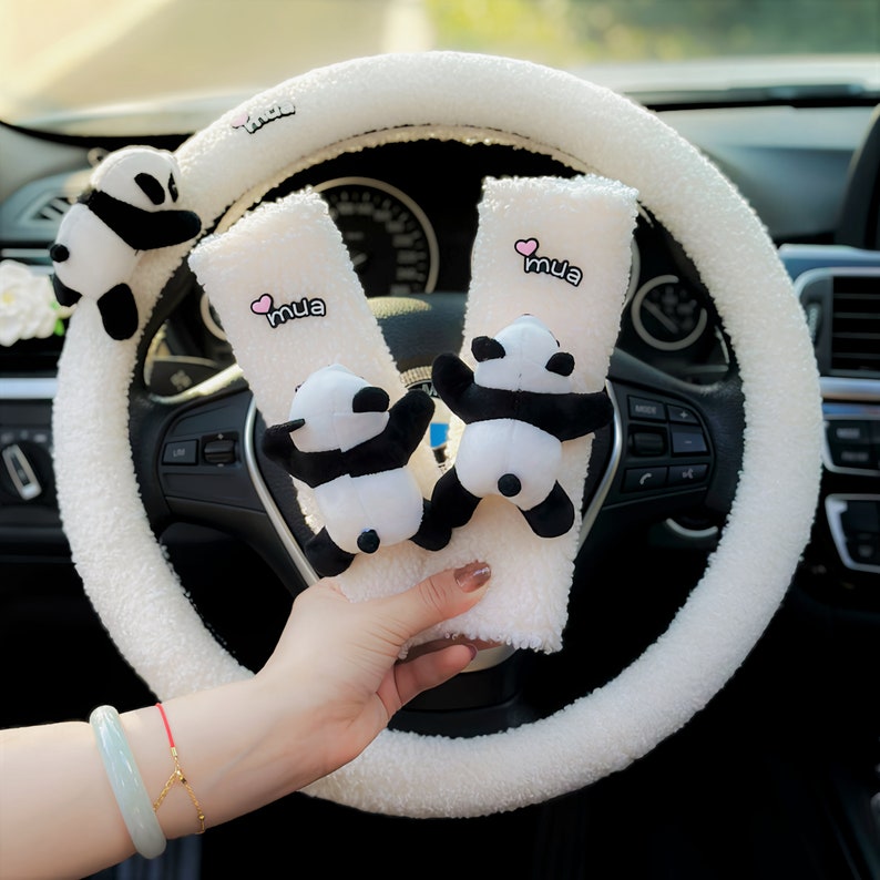May include: White fuzzy steering wheel cover with two plush panda bear seatbelt covers. The seatbelt covers have the word "mua" embroidered on them in pink.