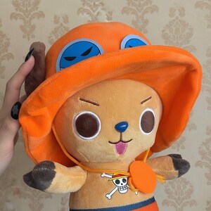 May include: A plush toy of a cartoon character with a large orange hat and blue goggles. The character has large brown eyes, a blue nose, and a pink tongue sticking out. The toy has a yellow necklace with a skull and crossbones emblem.