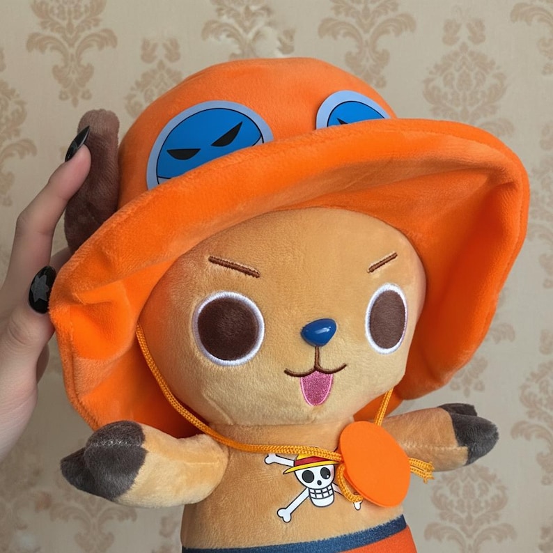 May include: A plush toy of a cartoon character with a large orange hat and blue goggles. The character has large brown eyes, a blue nose, and a pink tongue sticking out. The toy has a yellow necklace with a skull and crossbones emblem.