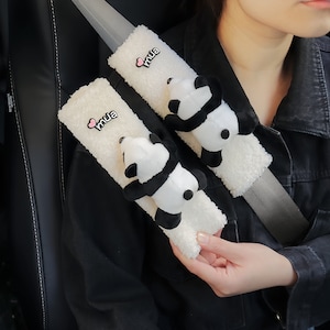 May include: Two white fuzzy car seatbelt covers with a panda bear plush attached to each. The covers have the word "mua" embroidered on them in black with a pink heart.