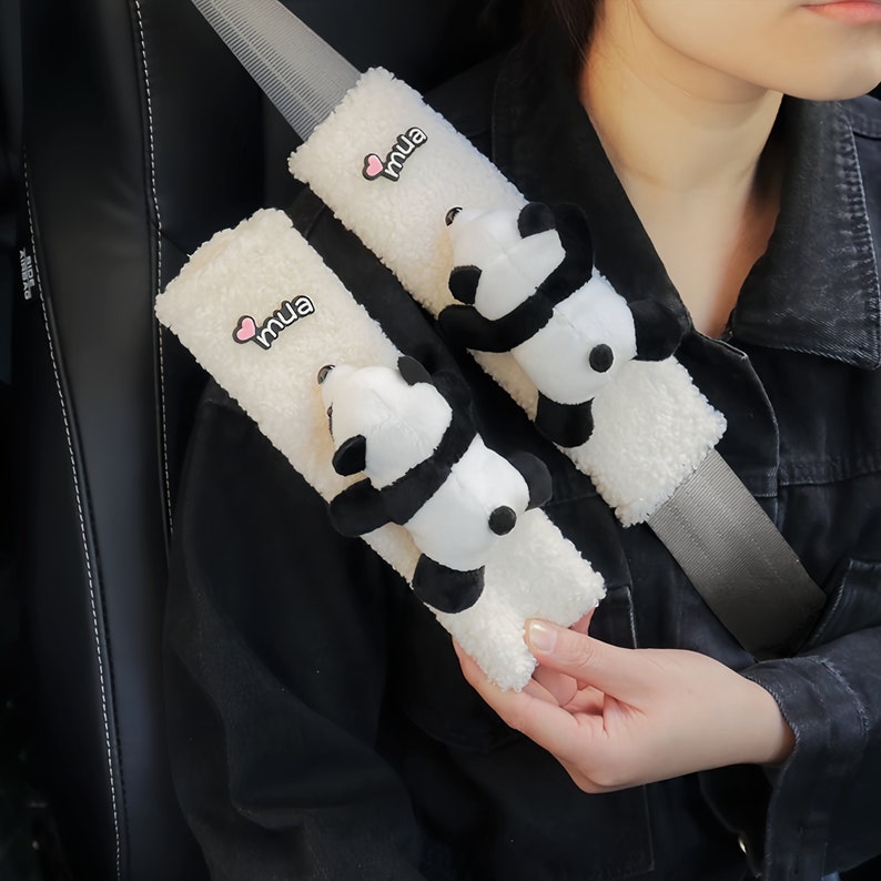 May include: Two white fuzzy car seatbelt covers with a panda bear plush attached to each. The covers have the word "mua" embroidered on them in black with a pink heart.
