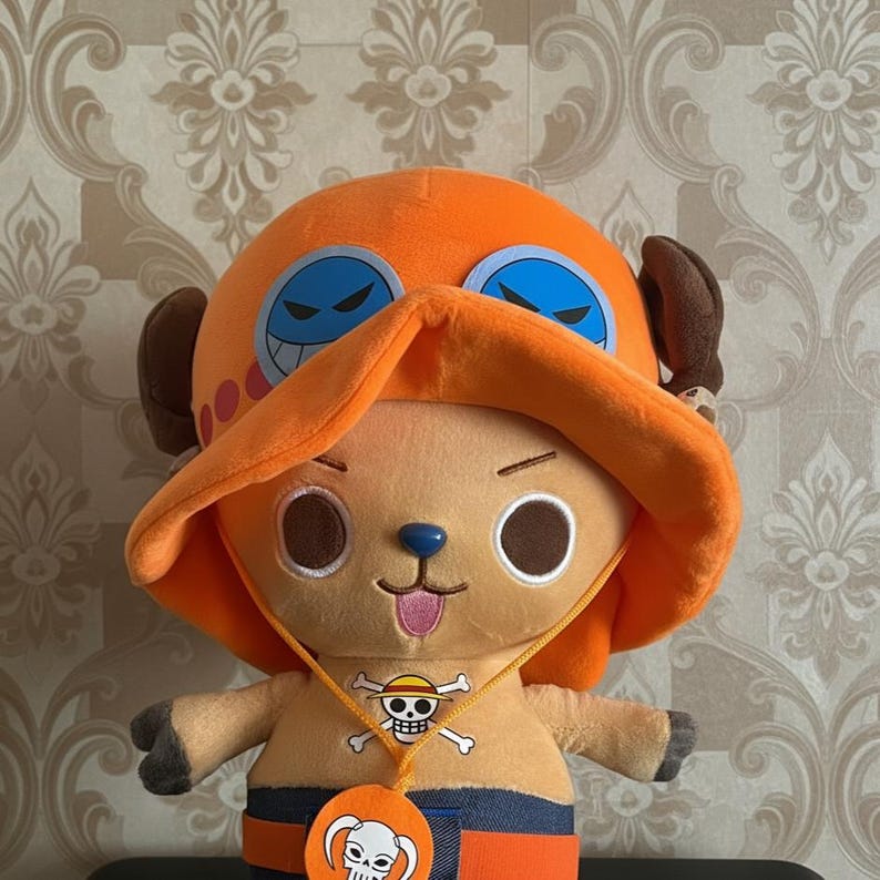 May include: A plush toy of a character with an orange hat and a blue nose. The toy has large, round eyes and a small, pink tongue sticking out. It wears a necklace with a skull and crossbones pendant. The character is from the anime series One Piece.