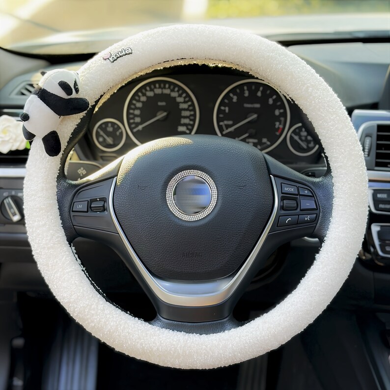 May include: White fluffy steering wheel cover with a panda bear charm attached. The cover has the word "vaiva" printed on it.
