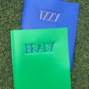 May include: Two personalised photo albums. One is bright blue with the name "IZZY" embroidered in white. The other is green with "BRADY" embroidered in blue. The albums are on a green grassy surface.