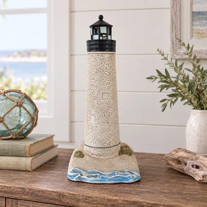 May include: A lighthouse figurine with a speckled beige tower, a black lantern room, and a base featuring textured sand and painted blue waves. The structure has small square windows and a conical roof, standing as a decorative nautical object.