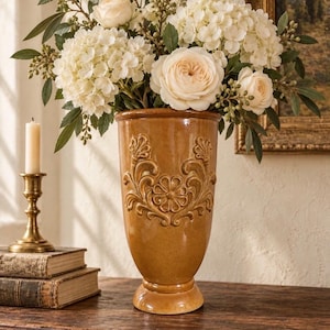 May include: A tall, glossy, amber-colored ceramic vase featuring an embossed floral scroll pattern on the front. The vase is filled with a large arrangement of white hydrangeas, pale cream roses, and green foliage with small berries.