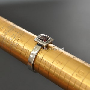May include: A silver ring with a square-shaped red gemstone set in a silver frame. The ring is on a sizing tool, showing a size 7.