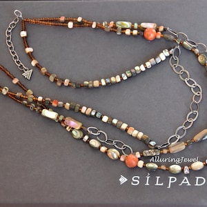 May include: A multi-strand necklace with brown, white, and gray beads, including shell and coral accents. The necklace features a silver chain and a small silver charm with a triangle design. The necklace is displayed on a gray background with the text "AlluringJewel" and "SÏLPADA" in white.