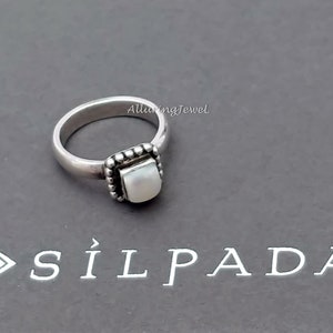 May include: A silver ring with a square white stone set in a beaded frame. The ring is on a gray surface with the text "Silpada" printed on it.