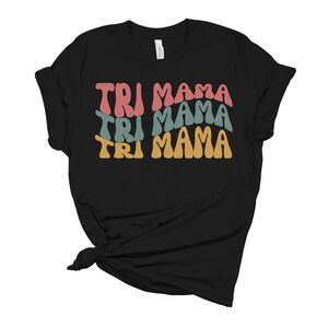 Op de afbeelding: Zwart T-shirt met de tekst "TRI MAMA" die drie keer wordt herhaald in een retro-lettertype. Elke "TRI MAMA" is in een andere kleur: roze, teal en geel.