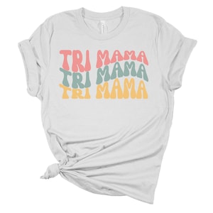 Op de afbeelding: Wit T-shirt met de tekst "TRI MAMA" die drie keer wordt herhaald in een retro-lettertype. De tekst is in tinten roze, blauw en geel.