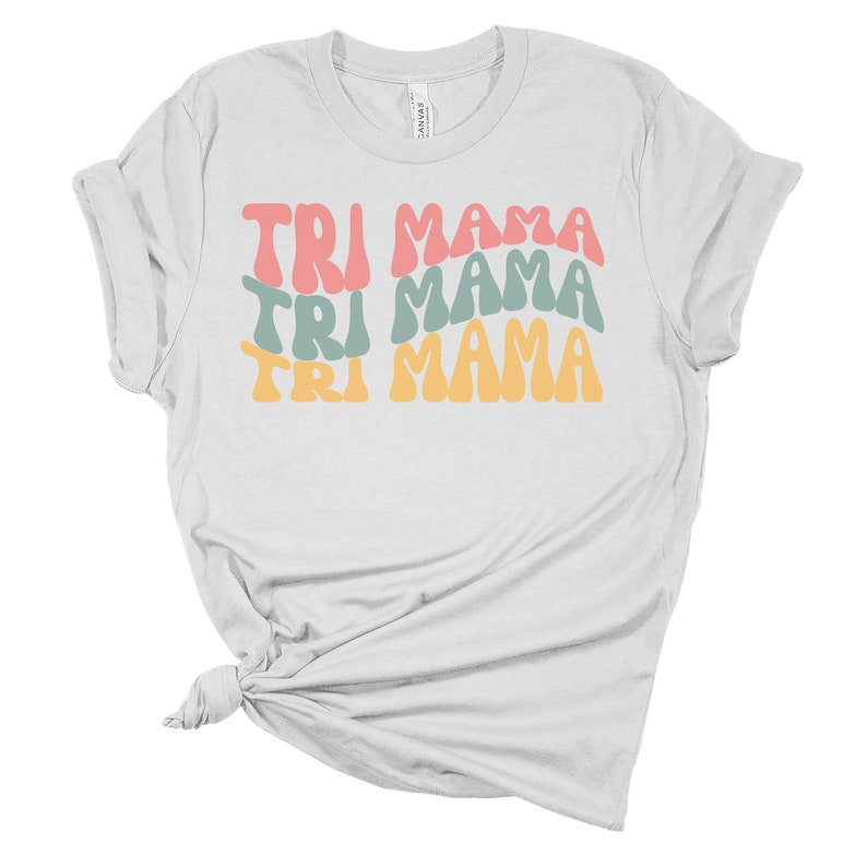 Op de afbeelding: Wit T-shirt met de tekst "TRI MAMA" die drie keer wordt herhaald in een retro-lettertype. De tekst is in tinten roze, blauw en geel.