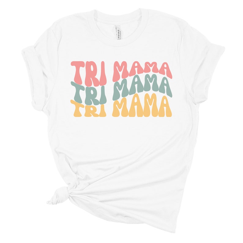 Op de afbeelding: Wit T-shirt met de tekst "TRI MAMA" die drie keer wordt herhaald in een retro-lettertype. De tekst is in tinten roze, groen en geel.