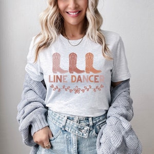 May include: A white t-shirt with a line dancing design. The design features three pink and orange cowboy boots with the text "LINE DANCER" below them. The design is surrounded by a floral border.