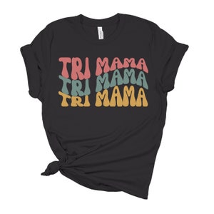 Op de afbeelding: Zwart T-shirt met de tekst "TRI MAMA" die drie keer wordt herhaald in een retro-lettertype. Elk woord heeft een andere kleur: roze, turquoise en geel.