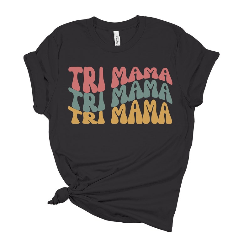 Op de afbeelding: Zwart T-shirt met de tekst "TRI MAMA" die drie keer wordt herhaald in een retro-lettertype. Elk woord heeft een andere kleur: roze, turquoise en geel.